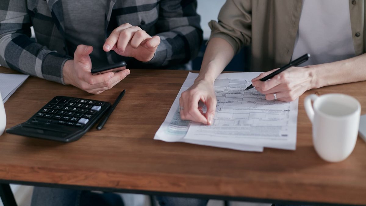 Duas pessoas preenchem formulários tributários e usam uma calculadora sobre uma mesa.
