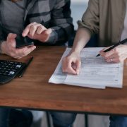 Duas pessoas preenchem formulários tributários e usam uma calculadora sobre uma mesa.