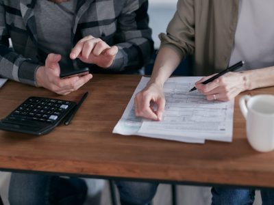 Duas pessoas preenchem formulários tributários e usam uma calculadora sobre uma mesa.