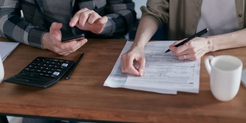 Duas pessoas preenchem formulários tributários e usam uma calculadora sobre uma mesa.