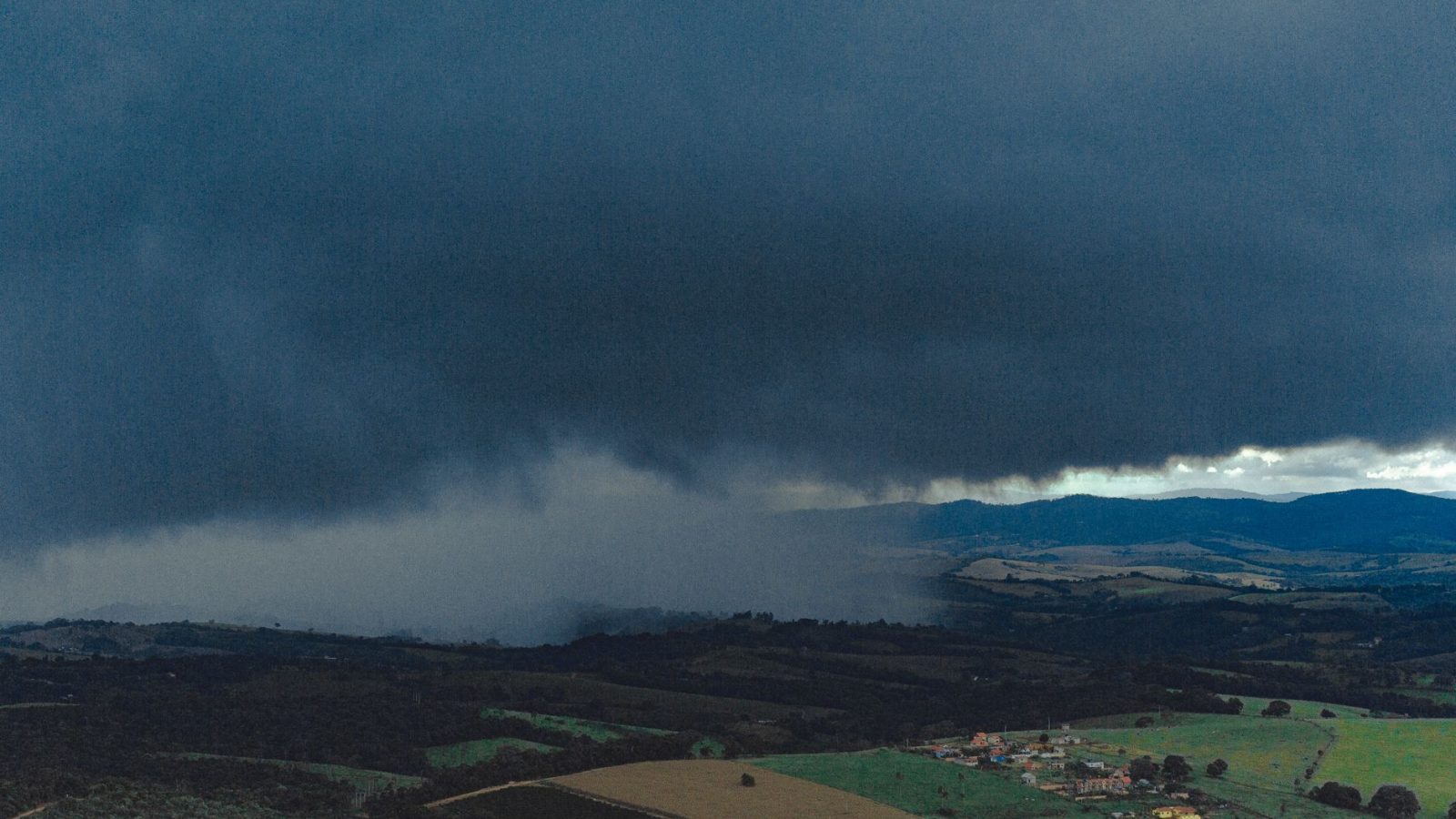 Vista aérea de extensas áreas agrícolas e montanhas ao fundo sob nuvens carregadas de chuva.