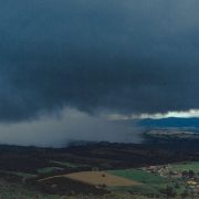 Vista aérea de extensas áreas agrícolas e montanhas ao fundo sob nuvens carregadas de chuva.