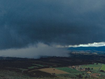 Vista aérea de extensas áreas agrícolas e montanhas ao fundo sob nuvens carregadas de chuva.