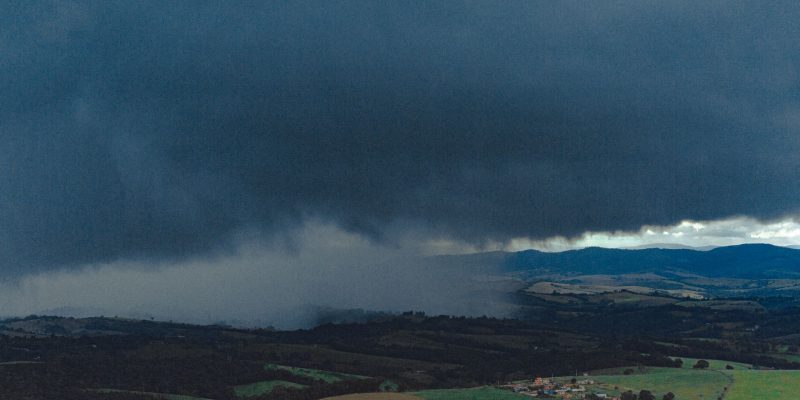 Vista aérea de extensas áreas agrícolas e montanhas ao fundo sob nuvens carregadas de chuva.