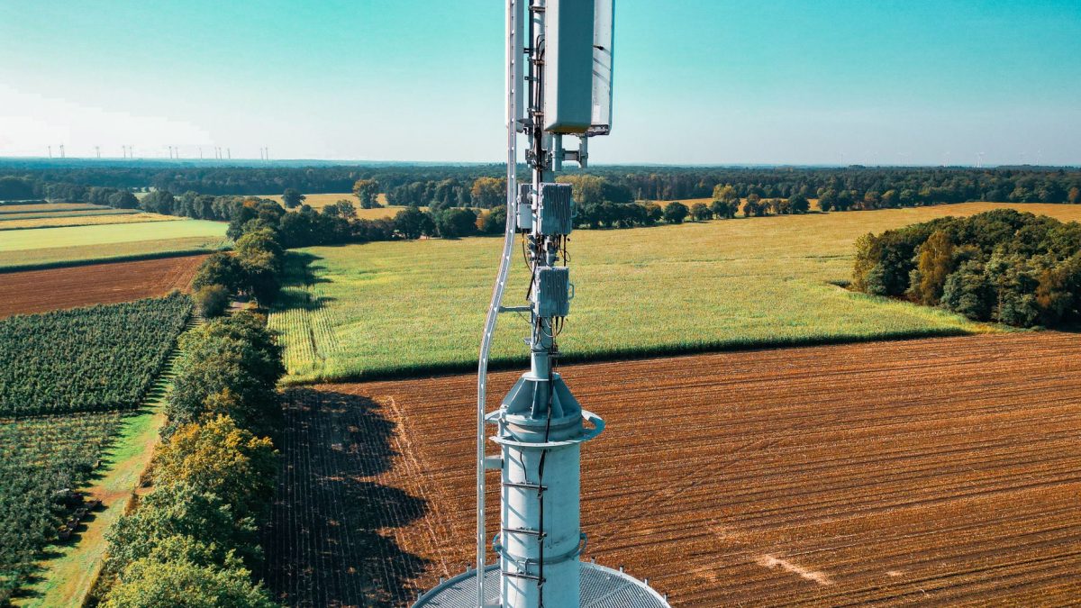 Torre de telefonia instalada em meio a campos agrícolas vistos de cima, cercados por áreas verdes.
