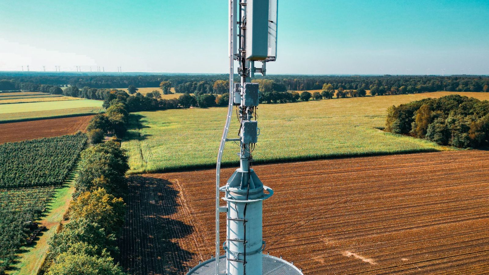 Torre de telefonia instalada em meio a campos agrícolas vistos de cima, cercados por áreas verdes.