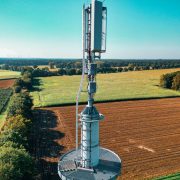 Torre de telefonia instalada em meio a campos agrícolas vistos de cima, cercados por áreas verdes.