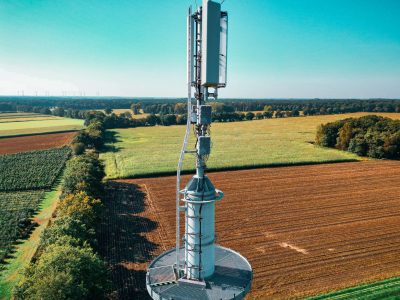 Torre de telefonia instalada em meio a campos agrícolas vistos de cima, cercados por áreas verdes.