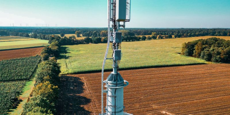 Torre de telefonia instalada em meio a campos agrícolas vistos de cima, cercados por áreas verdes.